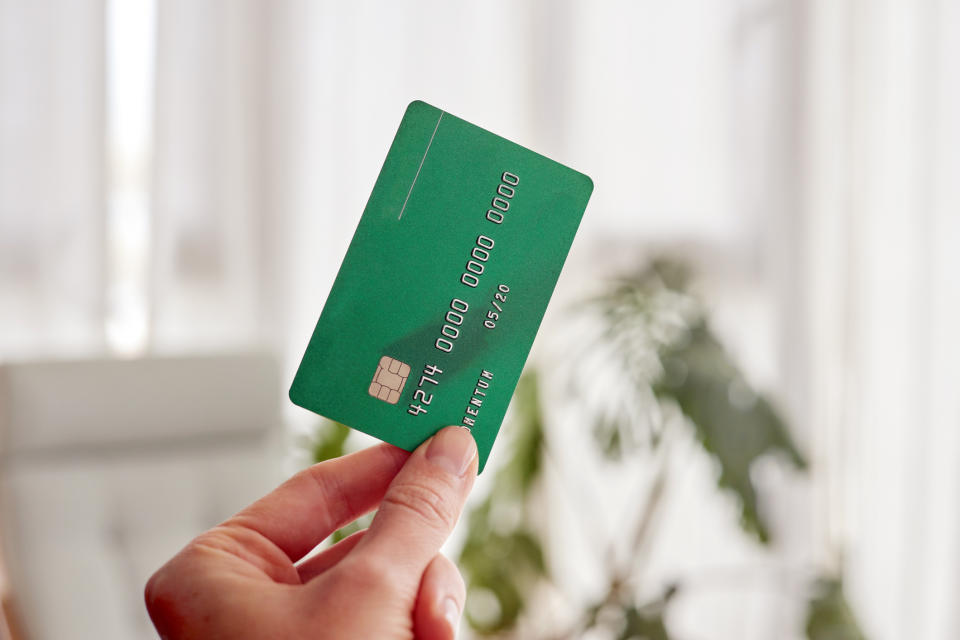 A hand holding a green credit card with the text "PREMIUM" visible, background shows blurred indoor plant and window