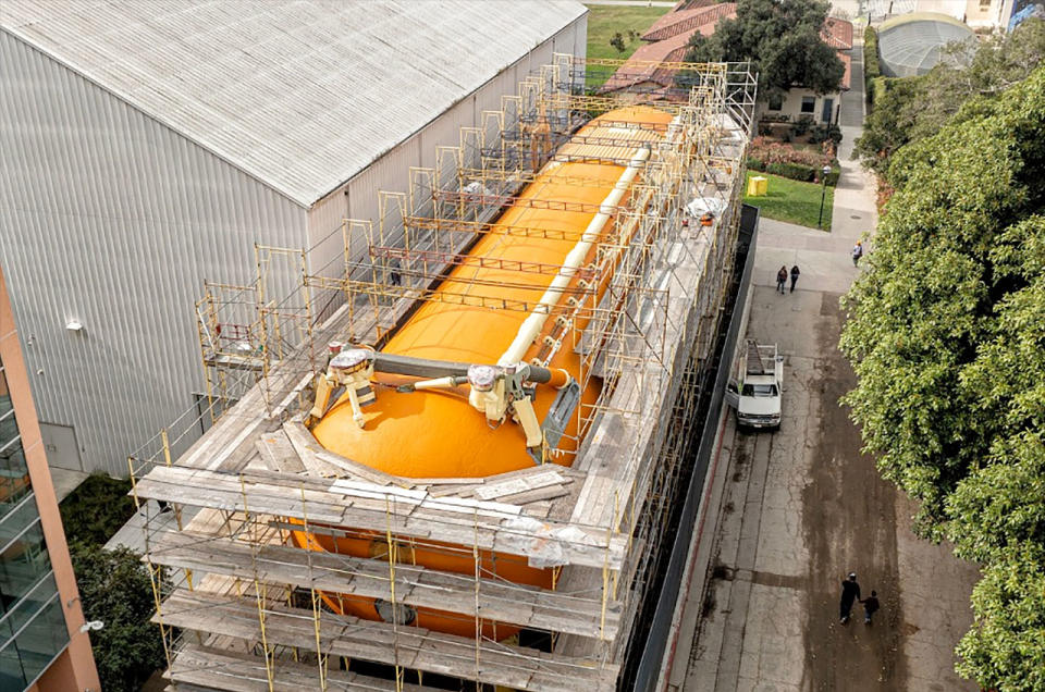 a space shuttle's orange external tank sits outside surrounded by scaffolding