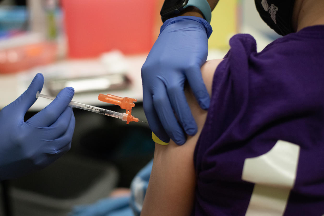 A health care worker wearing blue rubber gloves administers a a shot into the upper arm of someone wearing a face mask and purple T-shirt.