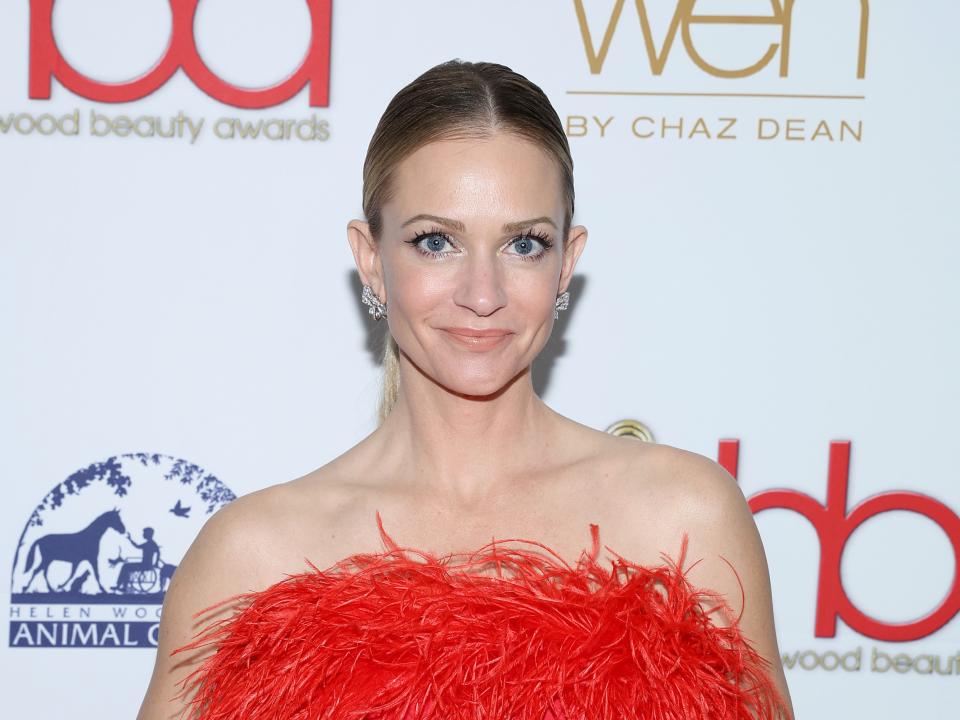 a.j. cook on a red carpet, wearing a form-fitting strapless red dress with a fringe at the top. she's smiling and holding a clutch