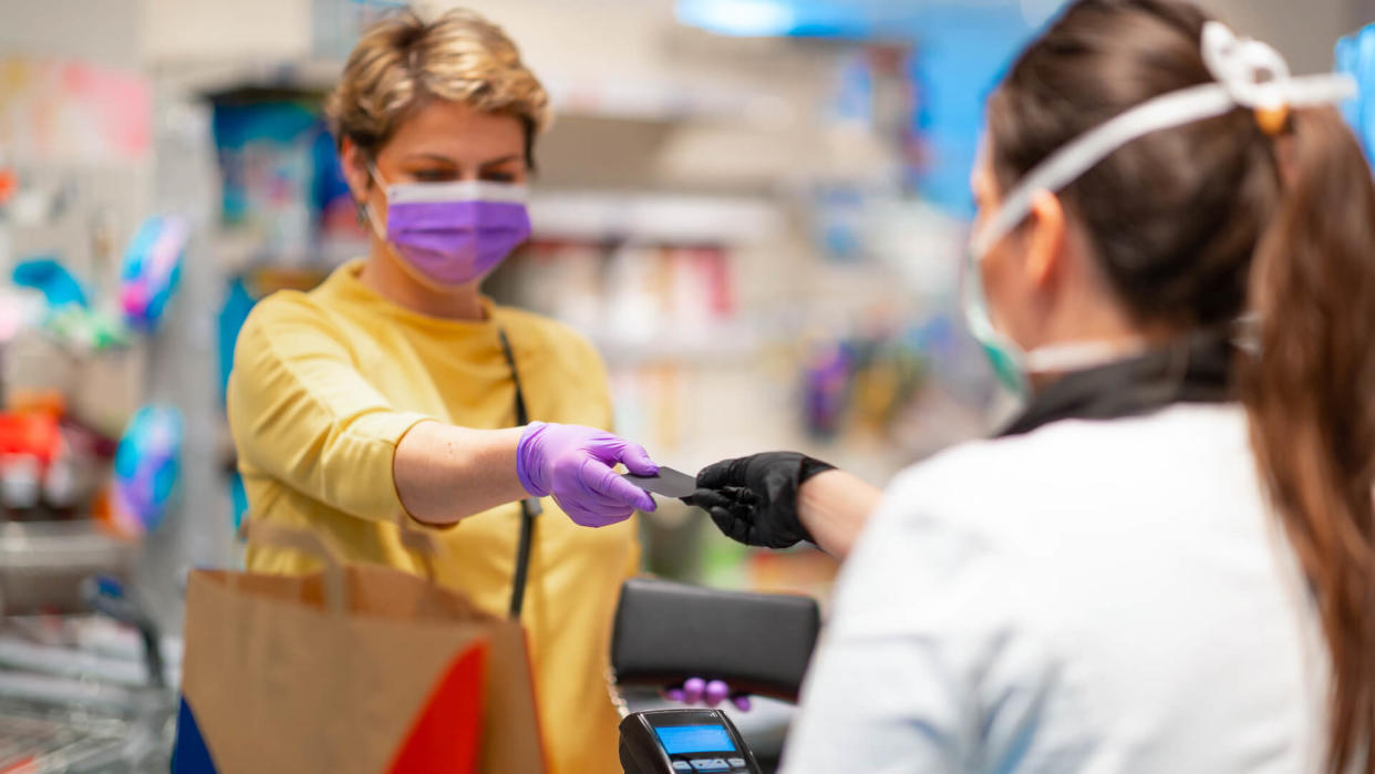 Cashier returning credit card at the cash register to woman with wallet wearing protective face mask and gloves to prevent viruses.