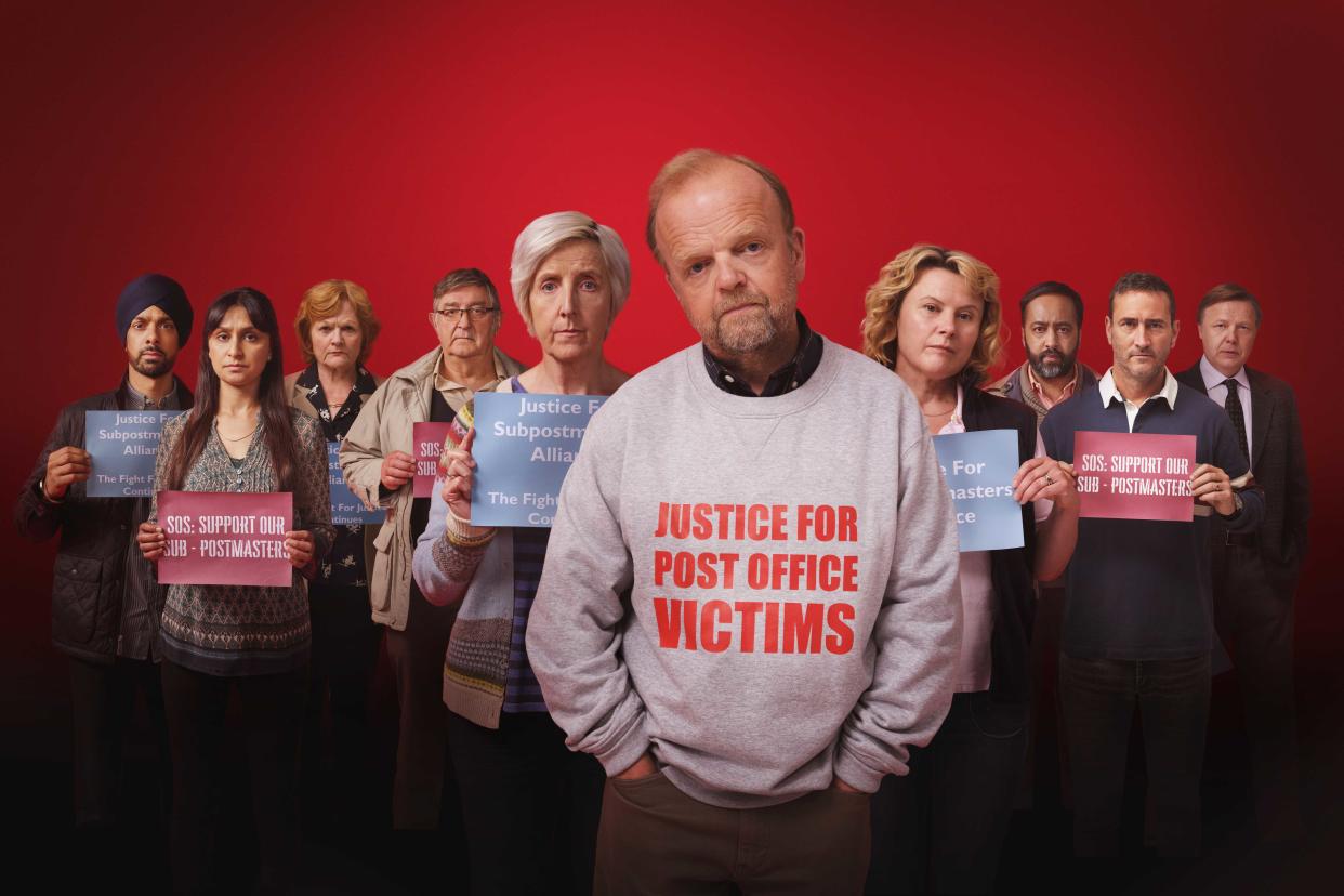 The cast of Mr Bates Vs The Post Office stand together holding signs against a red background.