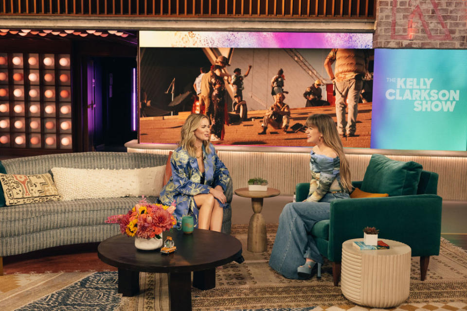 Kelly Clarkson interviews a guest on her show, both seated, with a screen showing a scene in the background
