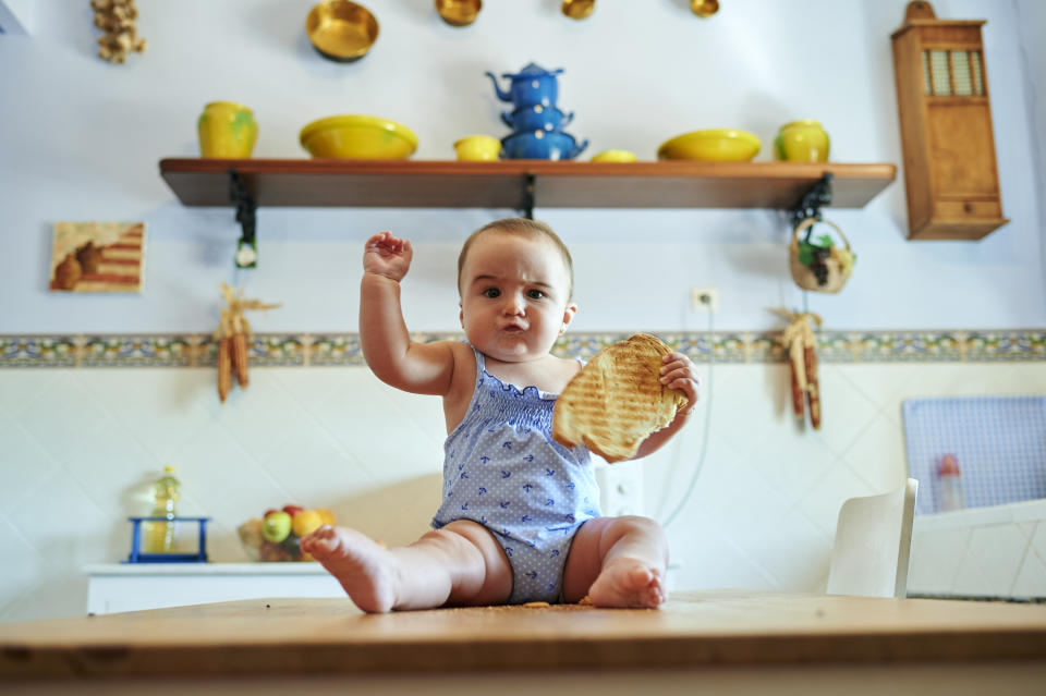 A baby sits on a kitchen table holding a slice of toast in one hand and holding his other fist up with a funny expression on his face