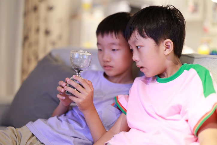 Two young boys are sitting on a couch, focusing on a glass object one of them is holding. Their expressions are curious and intent