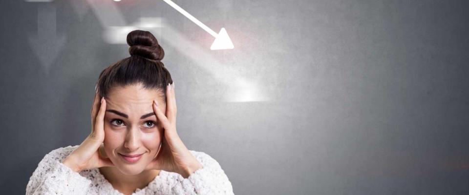 Stressed young businesswoman in white sweater sitting near concrete wall with falling graph drawn on it. Mock up