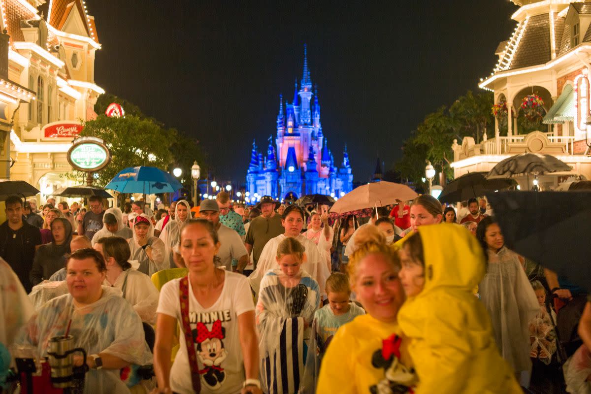 A large crowd of people walk towards the camera. A castle is lit up in blue behind them. On either side of the crowd are houses with lights strung up on them. 