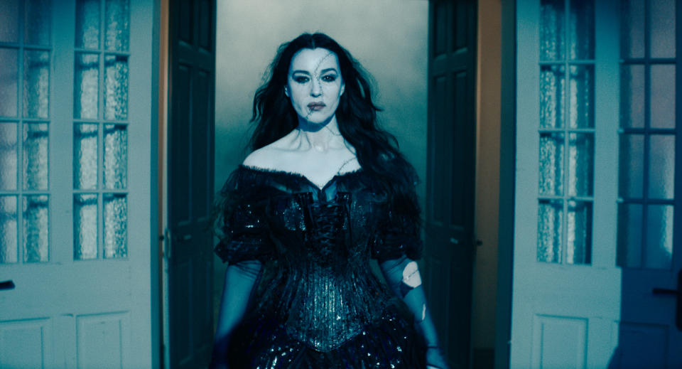 Monica Bellucci in a gothic-style outfit with dark, flowing hair stands in front of open doors in a dramatic, eerie setting