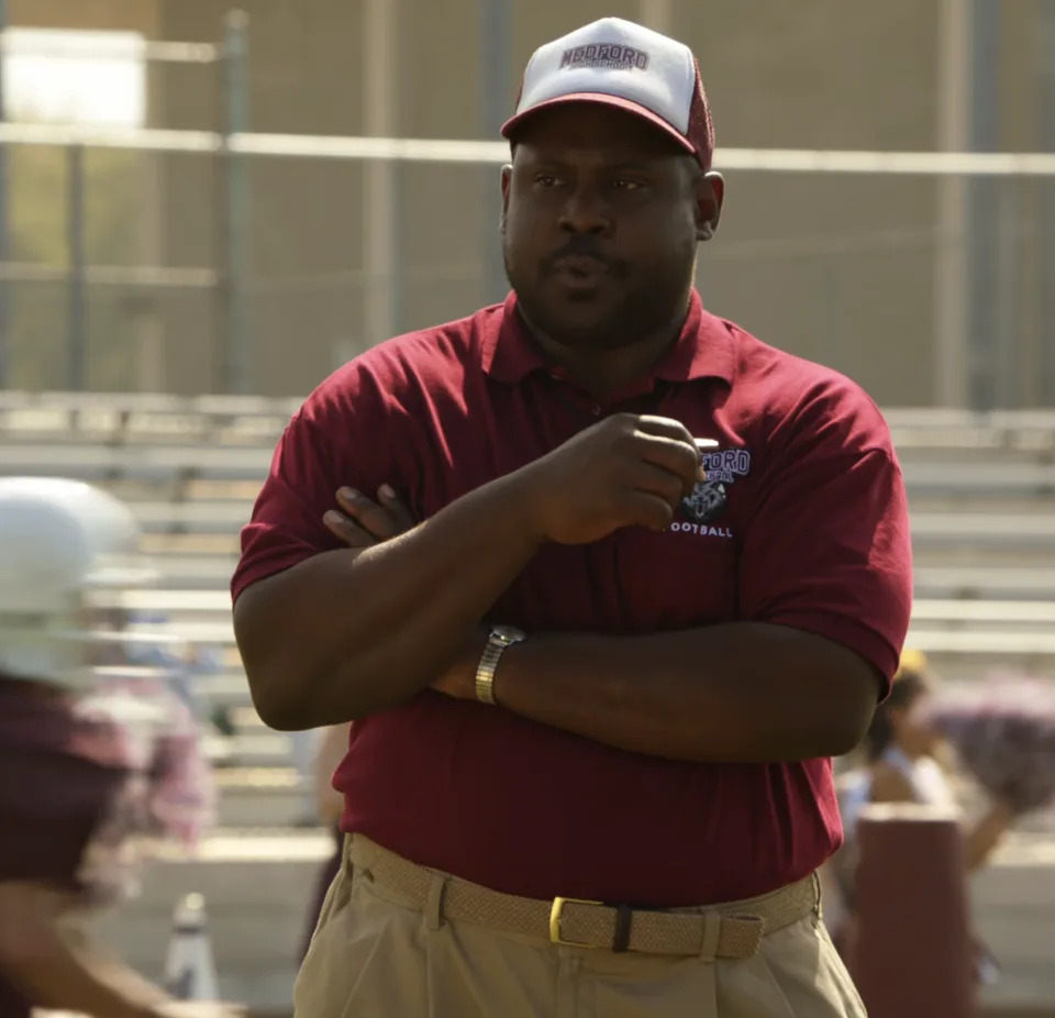 Wayne in a football coach attire with a whistle, on a practice field