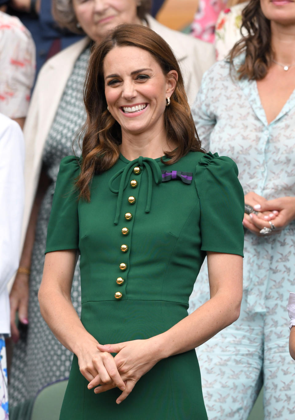 Catherine, Duchess of Cambridge in the Royal Box on Centre Court at Wimbledon in 2019 wearing a dark green, short-sleeved dress with gold buttons and ruffle details and a purple ribbon pinned to her left shoulder. Her long dark hair is loose.