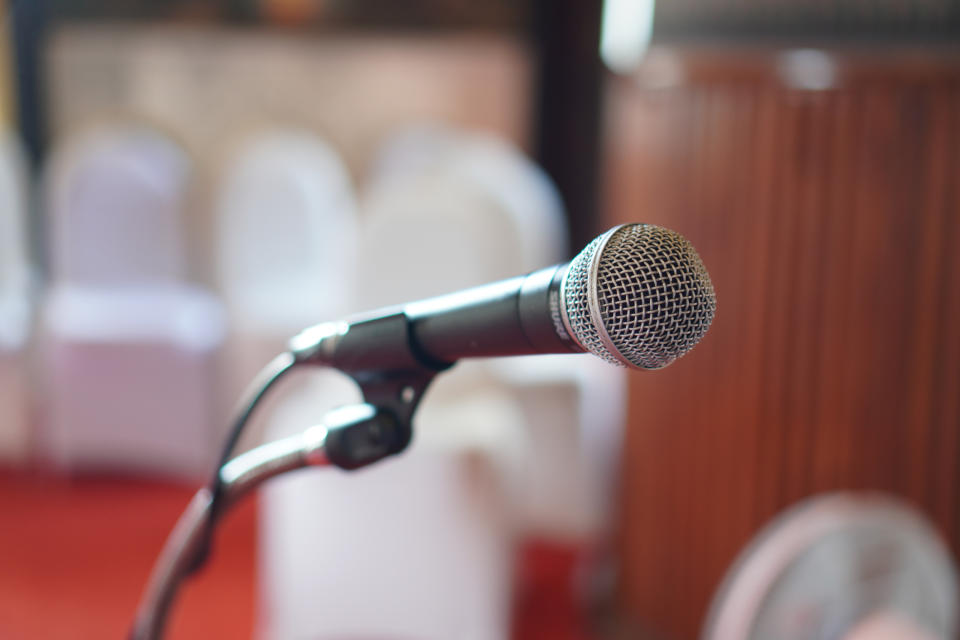 A microphone on a stand is the focal point, with blurred rows of empty wedding chairs in the background