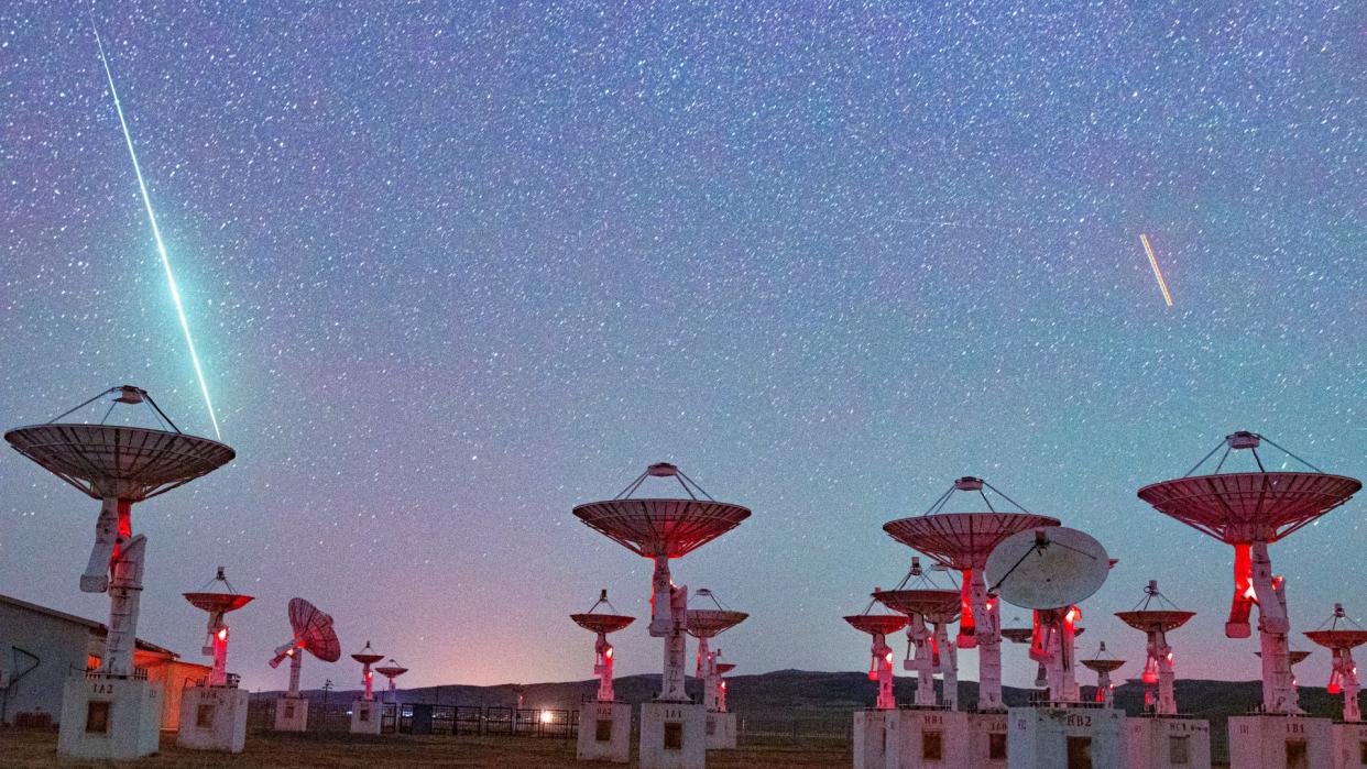  A green streak of light zooms among the stars above a set of antenna dishes in the desert. 