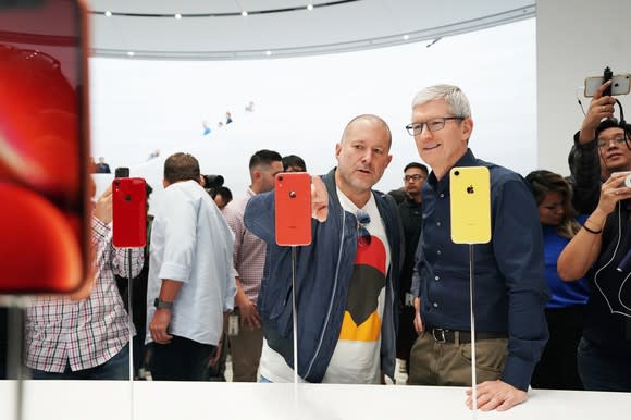 Apple executives Jony Ive and Tim Cook looking at red and yellow iPhone XR smartphones