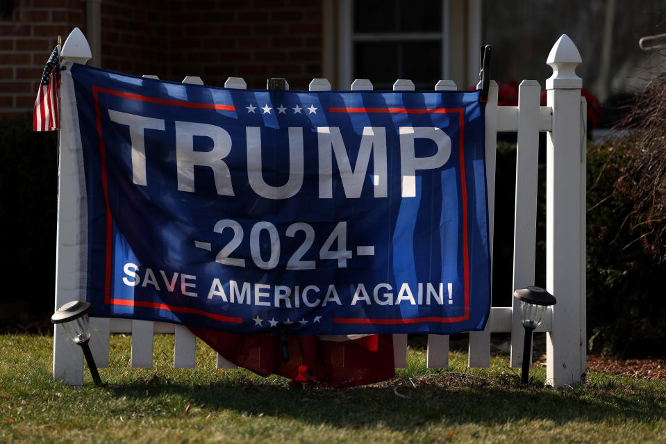 A banner hanging from a white fence says "TRUMP 2024 SAVES AMERICA AGAIN!"