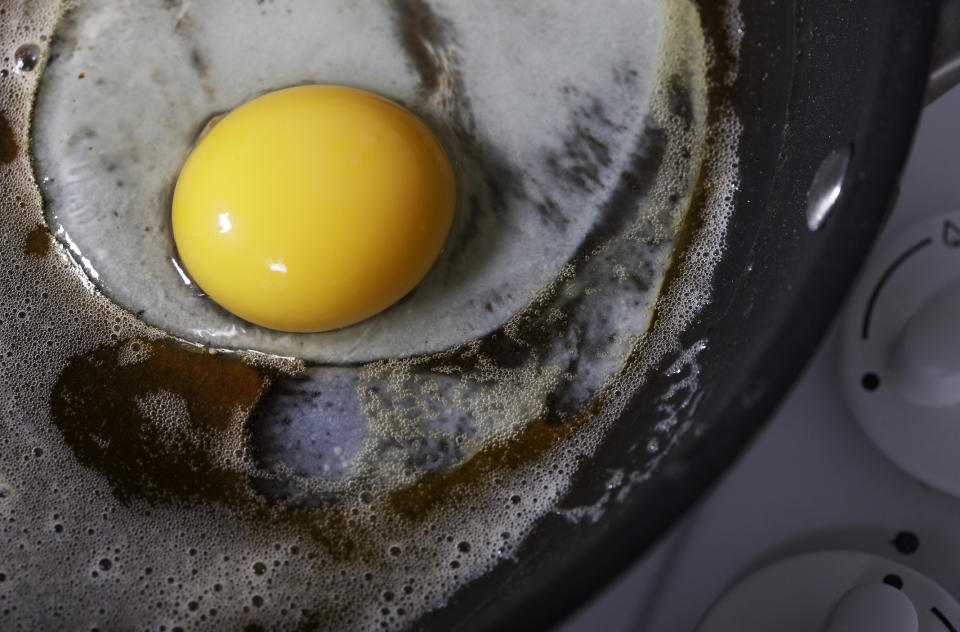 A raw egg is cracking into a buttered frying pan on a stove, ready to be cooked. The stove dials are partially visible