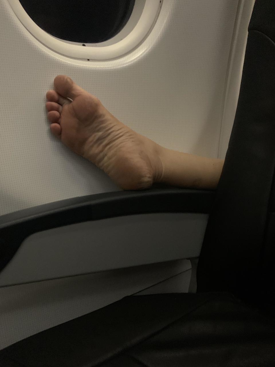 A foot is seen extended on the armrest of the seat in front on a plane. 