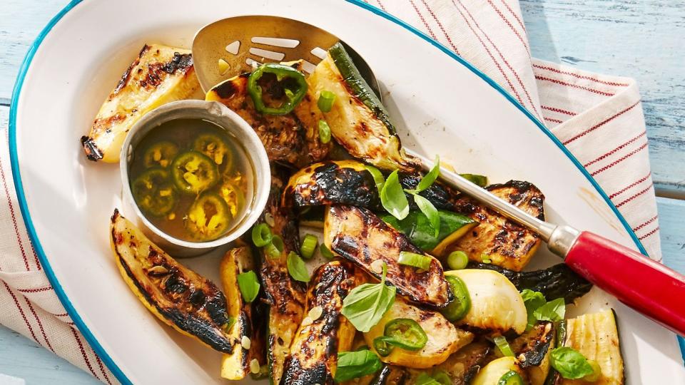 grilled summer squash in a white oval serving dish with a large spoon and a small bowl of lemon scallion dressing