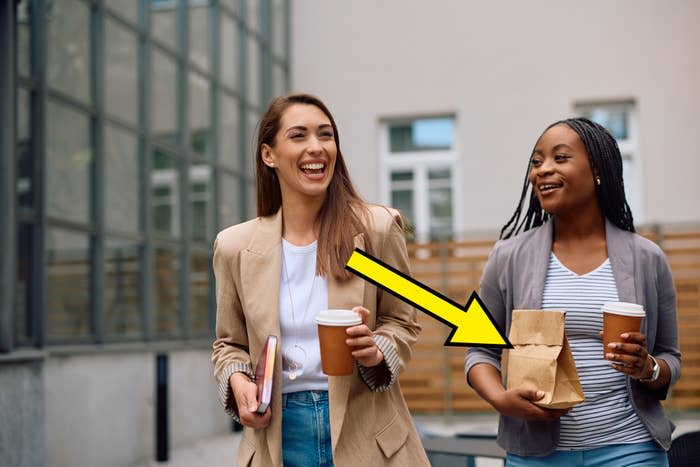 Two women, both casually dressed, are walking outside while smiling and holding coffee cups. One carries a notebook, and the other holds a brown paper bag