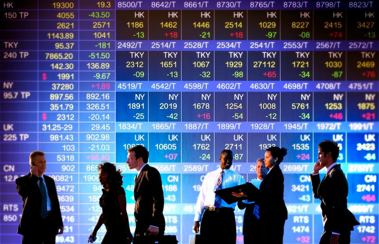 People in suits walk by and stand in front of a stock exchange presentation.