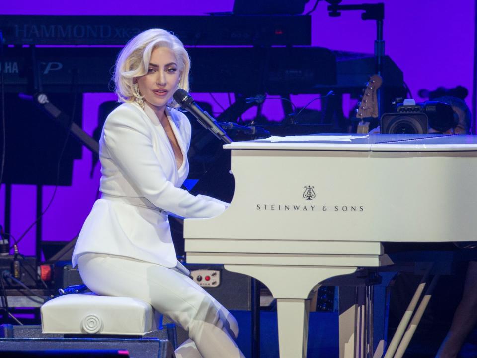 Gaga with a short blonde haircut in a white suit sitting and playing at a white piano.