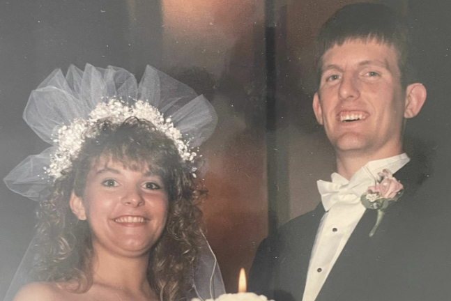 Bride and groom smiling behind wedding cake, bride in voluminous dress and veil, groom in tuxedo with bow tie