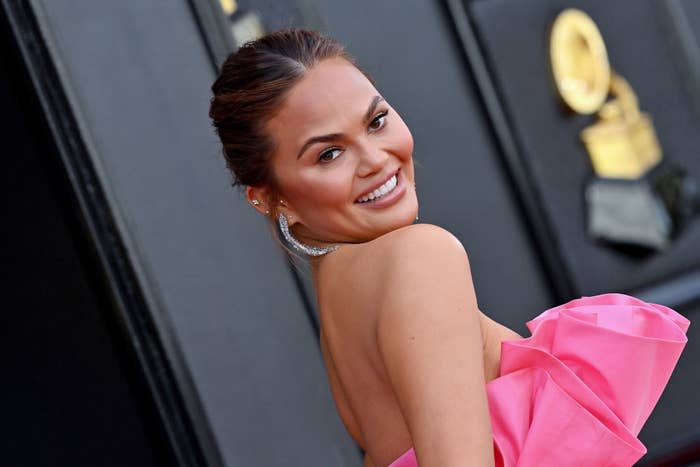 Chrissy Teigen smiling over her shoulder, wearing a strapless dress with a large ruffled detail