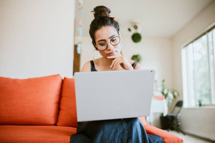 Woman with glasses and hair bun using a laptop on a couch, looking thoughtful