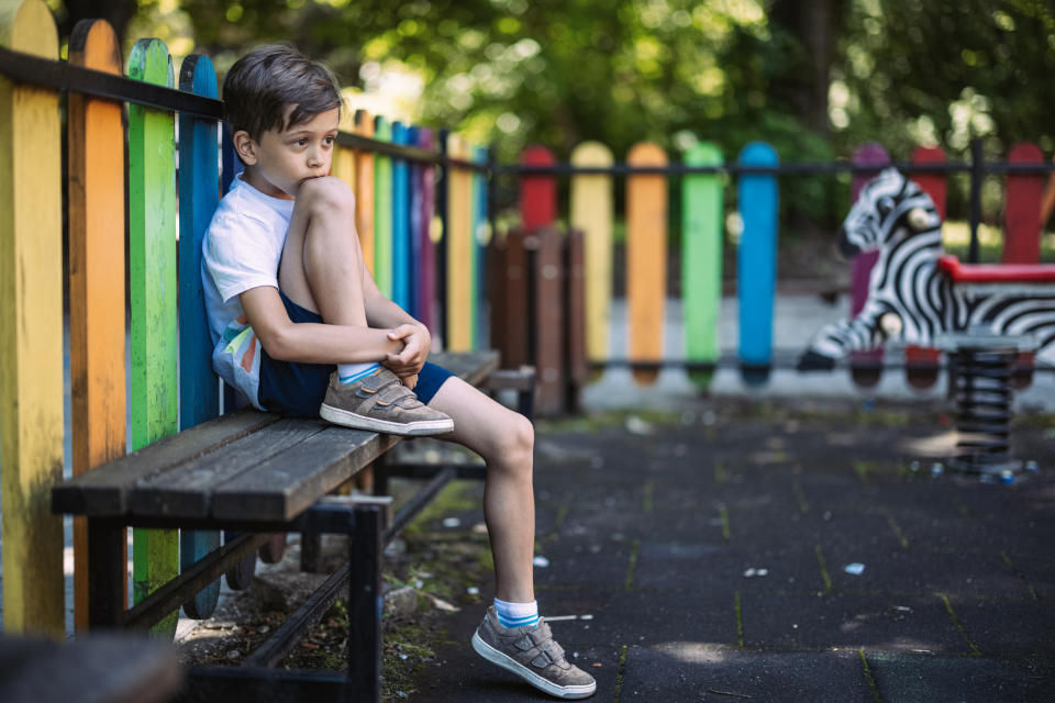 A boy sits on a bench in a playground, appearing thoughtful with his knee up to his chin. A colorful fence and a zebra spring rider are in the background