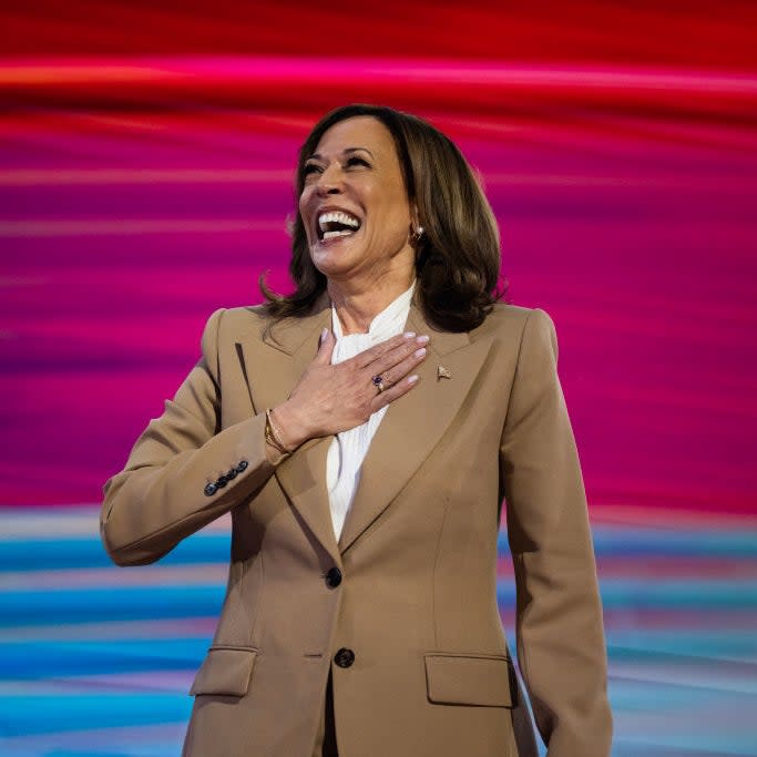 Kamala Harris is smiling and placing her hand on her chest while wearing a beige blazer and a white blouse. The background features colorful, wavy patterns