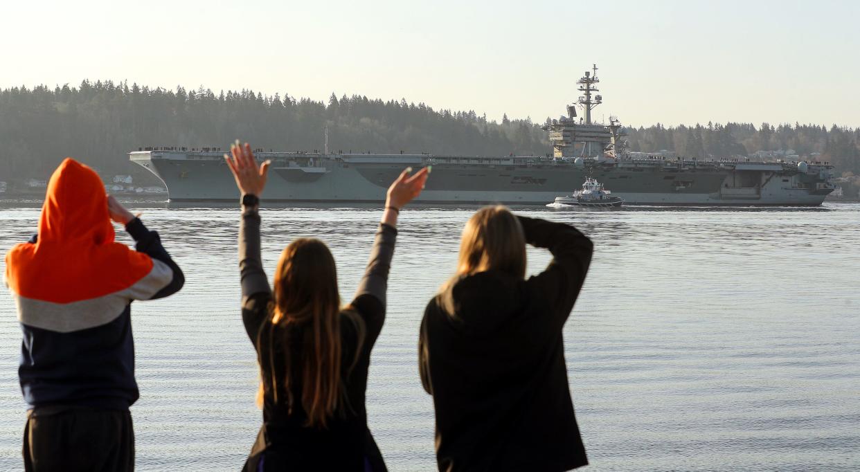 Family and friends of the sailors aboard the USS Theodore Roosevelt (CVN-71) wave from the shore of Bachmann Park in Bremerton, Wash. as the aircraft carrier departs Puget Sound Naval Shipyard after its 18-month overhaul, on Friday, March 17, 2023.