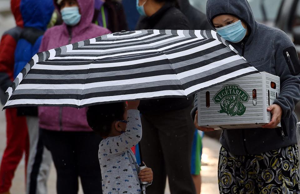 A small child holds a big umbrella while their mom in a mask holds a box that says 'vegetables.'