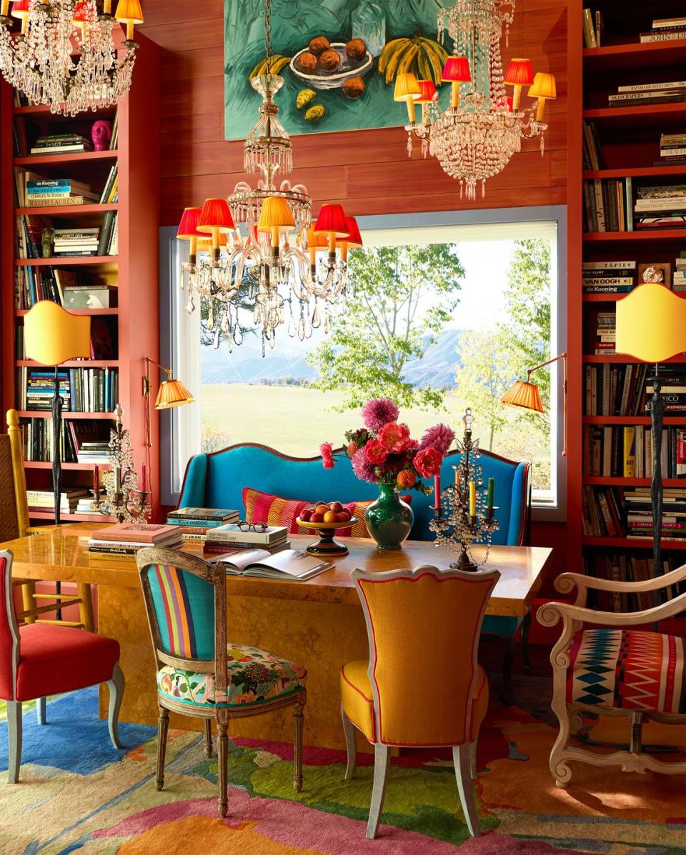 dining room table with mismatched cushioned chairs and a bright blue settee, flowers and candelabra on marble table, painting above window and sconces and bookshelves on both sides, three chandeliers