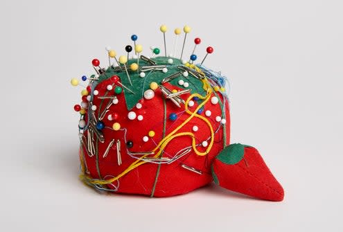 A pincushion shaped like a tomato, filled with various colored pins and tangled threads, is displayed against a plain background