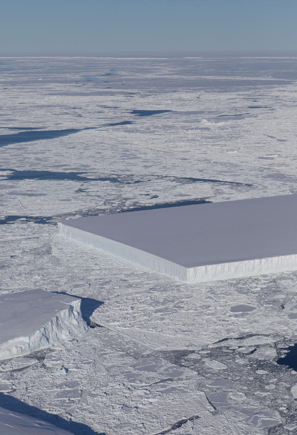 Aerial view of a large rectangular iceberg floating among irregularly shaped icebergs in a vast frozen landscape