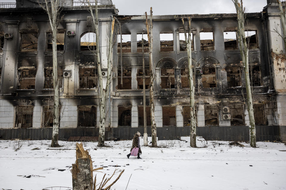 A person with a pink bag walking in the snow passes by a destroyed building.