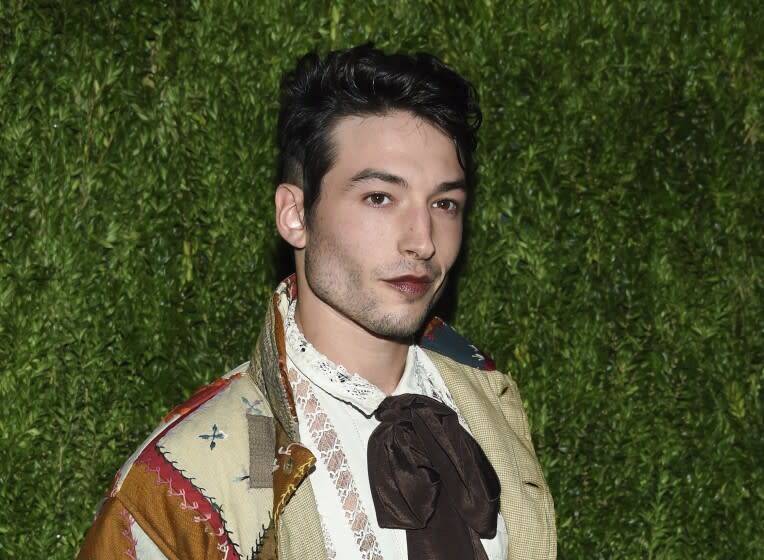 A person with short dark hair, stubbly facial hair and dark lipstick poses in front of foliage