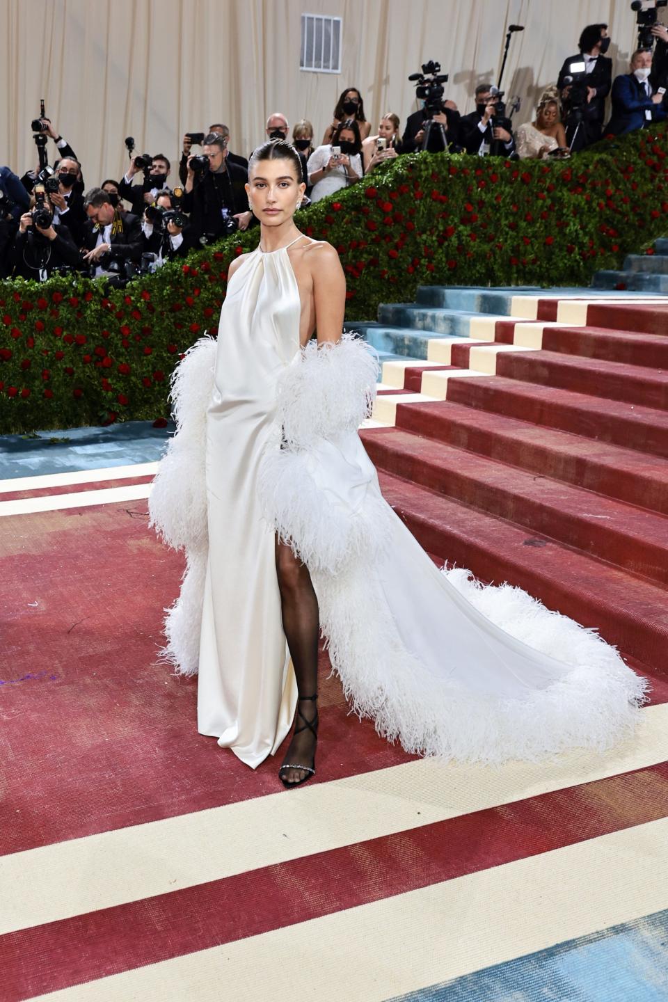 Hailey in an off-white silk halter top dress with an open back. The dress fabric ruches at her hip and has a leg slit that reveals black sheer stockings and black heels. She's wearing an oversized white cape off her shoulders lined with white feathers on the arms and hemline.