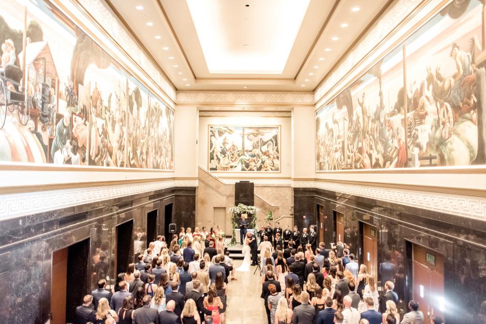 A wedding is conducted inside IU Auditorium's Hall of Murals, with the Thomas Hart Benton murals.