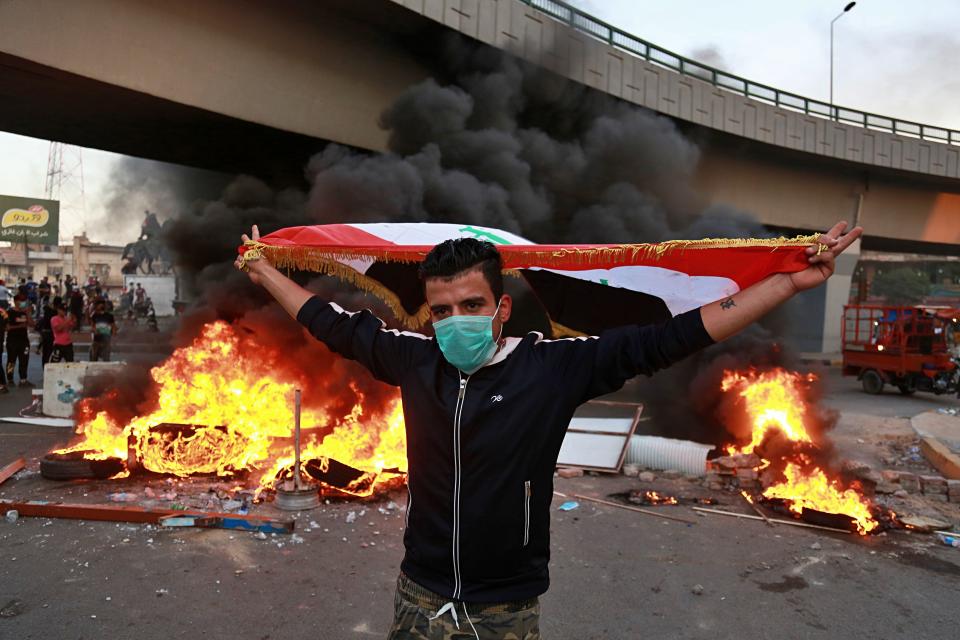 A protester holds an Iraqi flag while anti-government protests set fire while security forces fired live ammunition and tear gas near the state-run TV in Baghdad, Iraq, Monday, Nov. 4, 2019. (AP Photo/Khalid Mohammed)