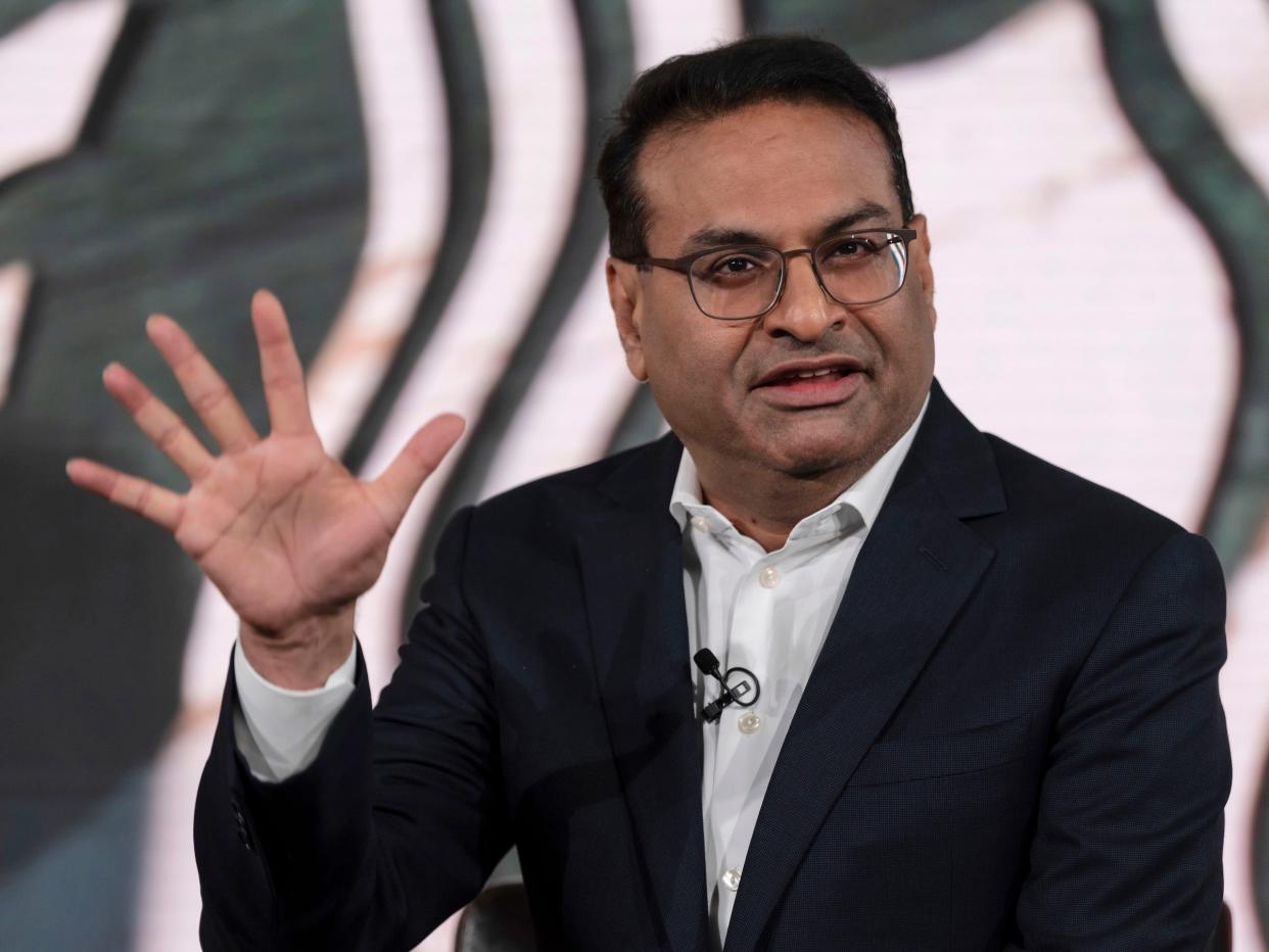 Laxman Narasimhan holds up his hand while speaking onstage in front of a large Starbucks logo.