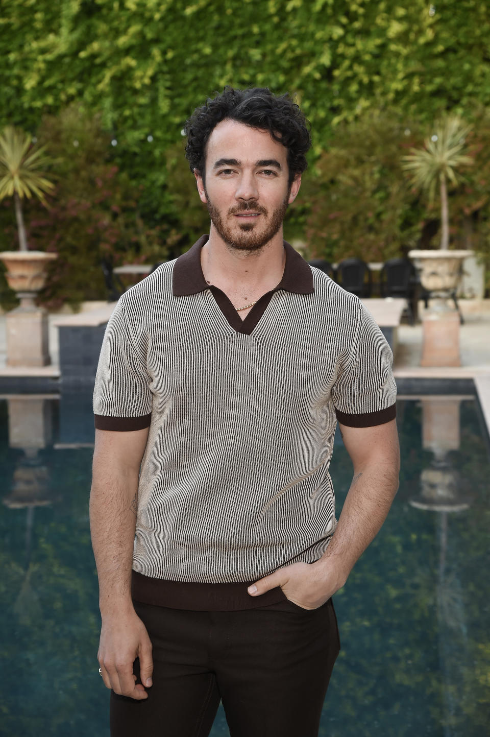 Kevin Jonas stands in front of a pool and greenery, wearing a vintage-style short-sleeve shirt with dark trim and matching pants