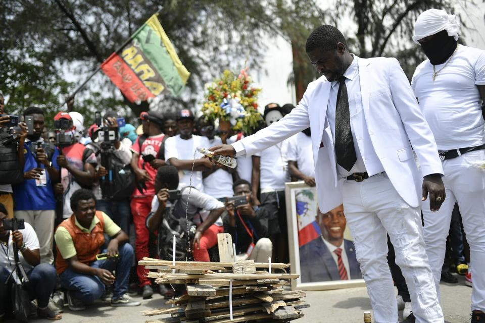 A man in a white suit and black tie pours liquid from a bottle onto a pile of wooden planks as a crowd watches