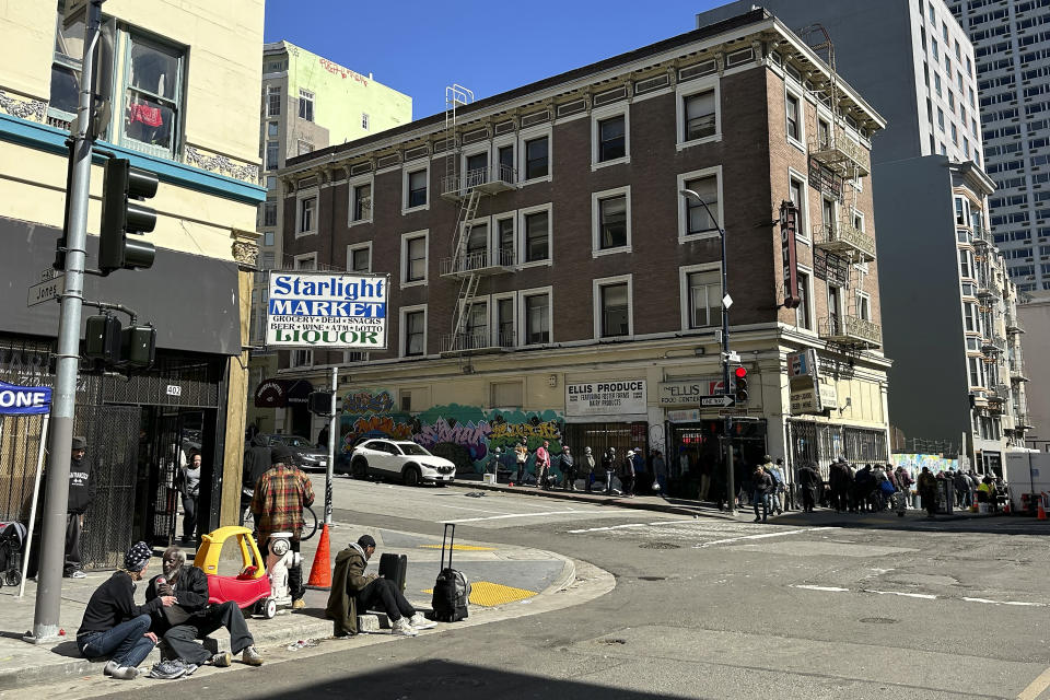 People sit on the curb at the corner of Ellis Street and Jones Street in the Tenderloin neighborhood of San Francisco, Thursday, March 14, 2024. Two hotels and unnamed residents of one of San Francisco's most problematic neighborhoods for drug use and tent encampments sued the city Thursday, demanding that city officials stop using the district as a containment zone. (AP Photo/Jeff Chiu)