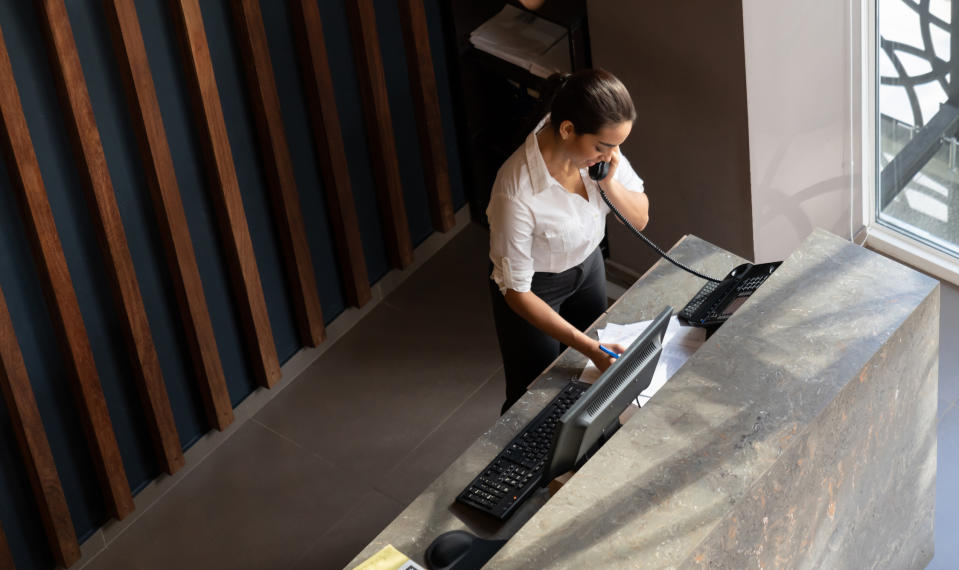 A woman in business attire talks on the phone at a reception desk, working with a computer and paperwork