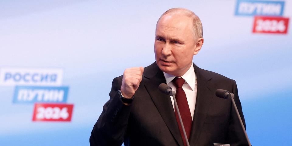 Russian President Vladimir Putin, wearing a dark suit and red tie, holds a clenched fist up against a blue background with writing advertising Russia's election.