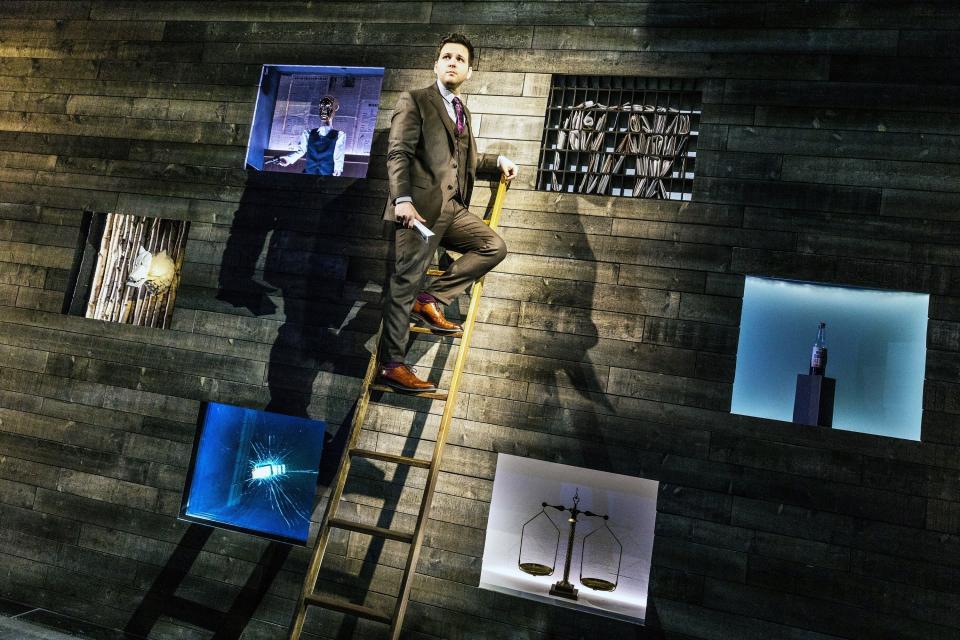 A white man in a suit stands on a ladder leading against a wooden wall on a stage. Surrounding him are six boxes open in the wall with various items - a bottle, a scale, broken glass, a fox, a puppet, and mail.