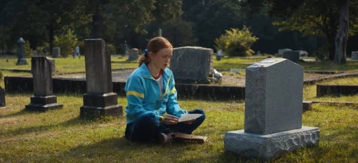 Max, a young girl with red hair tied back in a ponytail, holds a letter while sitting in front of a gravestone.  She wears a blue zip up hoodie with a yellow horizontal stripe across the upper arm, and blue jeans.