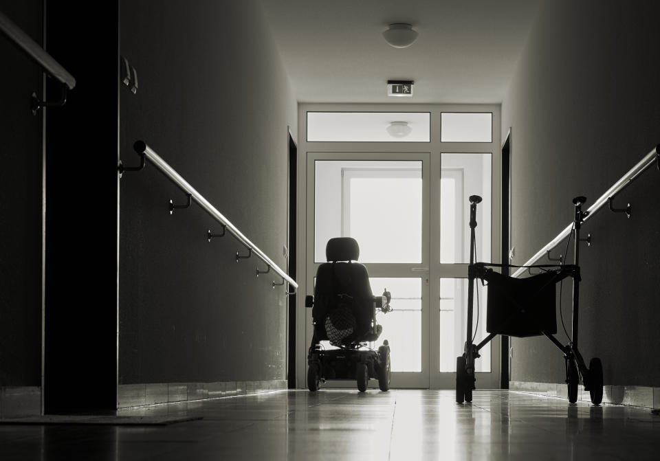 Person in wheelchair at the end of a hallway, facing a bright doorway, with a walker on the right side