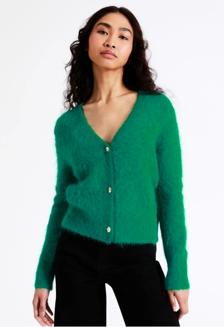 A model with long dark hair wears a bright green cardigan in fluffy wool with white buttons, over black pants.