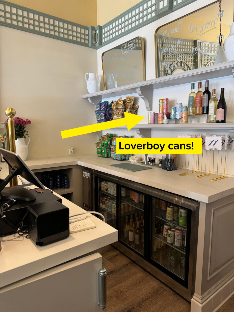 A small café counter with shelves displaying snacks, drinks, and wine bottles. A cash register and small order number holders are on the counter